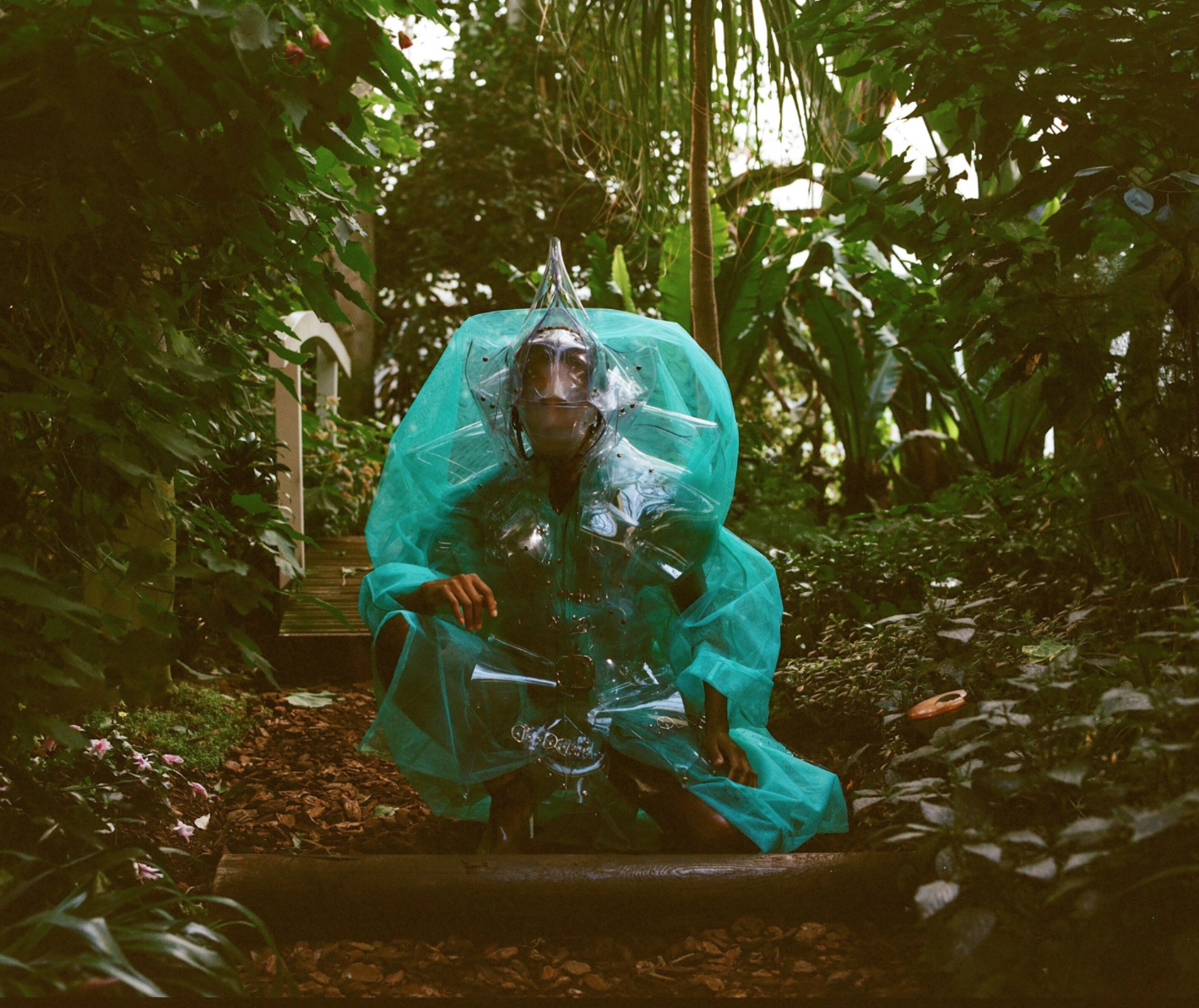 A photograph of a person dressed with an alien mask wearing puffy translucent green garbage bags that also bubble up over a device strapped to their back. They squat in the center framed by a lush green forest.