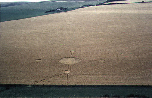 An aerial photograph of a crop circle in a light brown field. The crop circle looks like a large circle with 4 perpendicular lines coming out of it, at the end of each line is a smaller circle, at the bottom small circle are two lines that form a triangle.