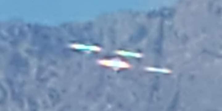 A blurry color photograph of a cluster of 4 blurry oblong opalescent objects moving in front of a mountain range. The top right triangle of the frame is a deep blue sky.