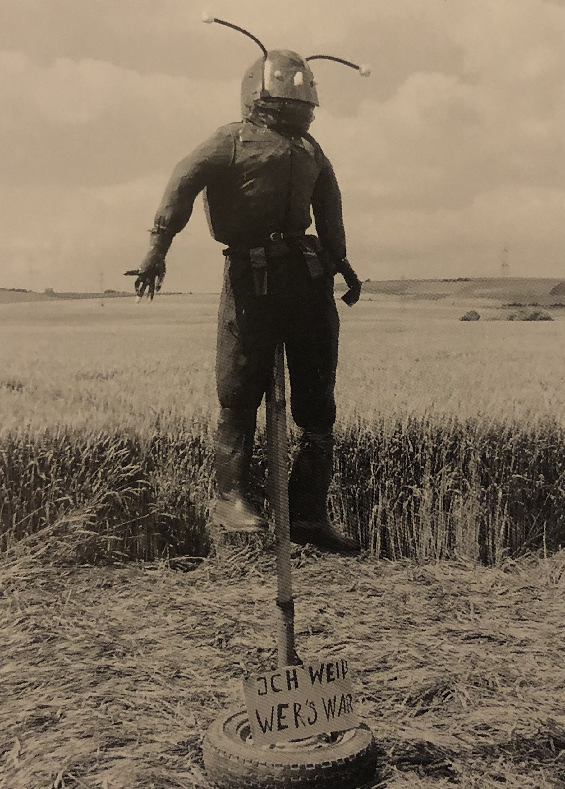 A sepia photograph of a scarecrow dressed as an alien is positioned standing, held up by a wooden pole sticking out of a tire sitting in a flattened part of a wheat field in the foreground with a line of upright wheat behind it and the field in the background. The horizon line cuts horizontally ⅔ of the way up the frame with small hills and power lines visible. The top half of the alien figure looms in the sky with gnarled glove hands and a head made of a helmet with eyes and a nose glued on and 2 antennae with small balls at the end sticking out of the top. A hand painted sign sits atop the tire at the bottom of the wooden stake, reading 'ICH WEIß, WER’S WAR'.