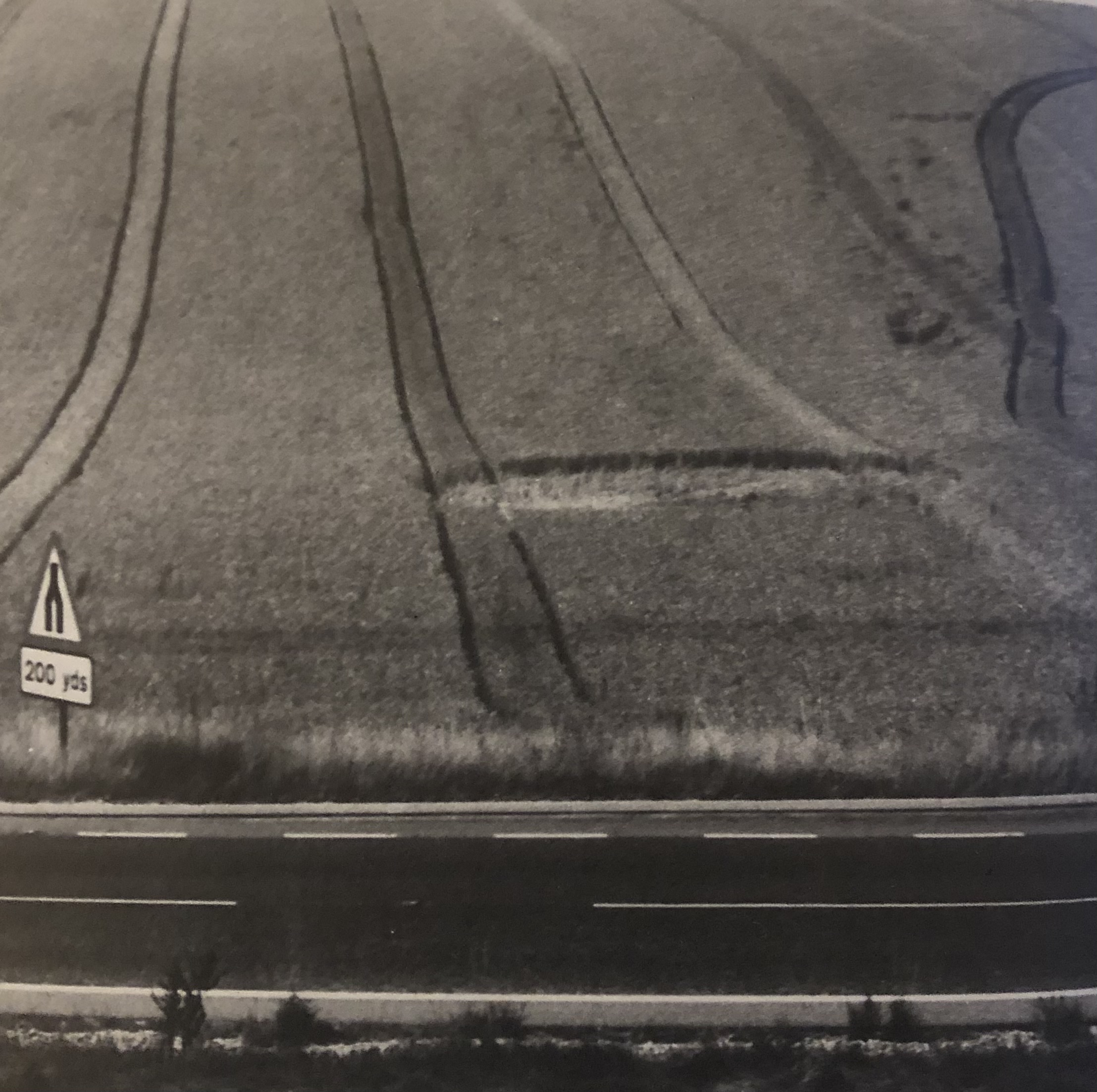 A black and white side-aerial photograph where the ellipse of a crop circle can be seen in a field beyond a paved road and road sign at the bottom of the frame. Parallel lines follow the topography of the field down from the top of the frame to meet the grass that lines the road.